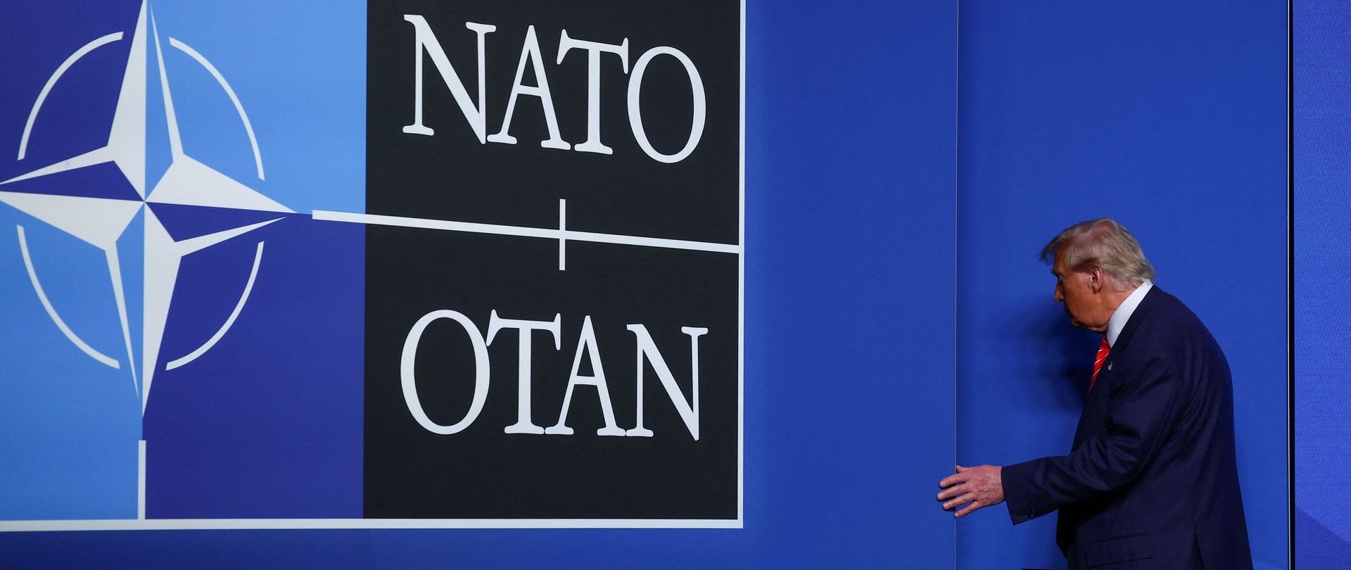 Donald Trump next to a blue wall with the NATO logo on it