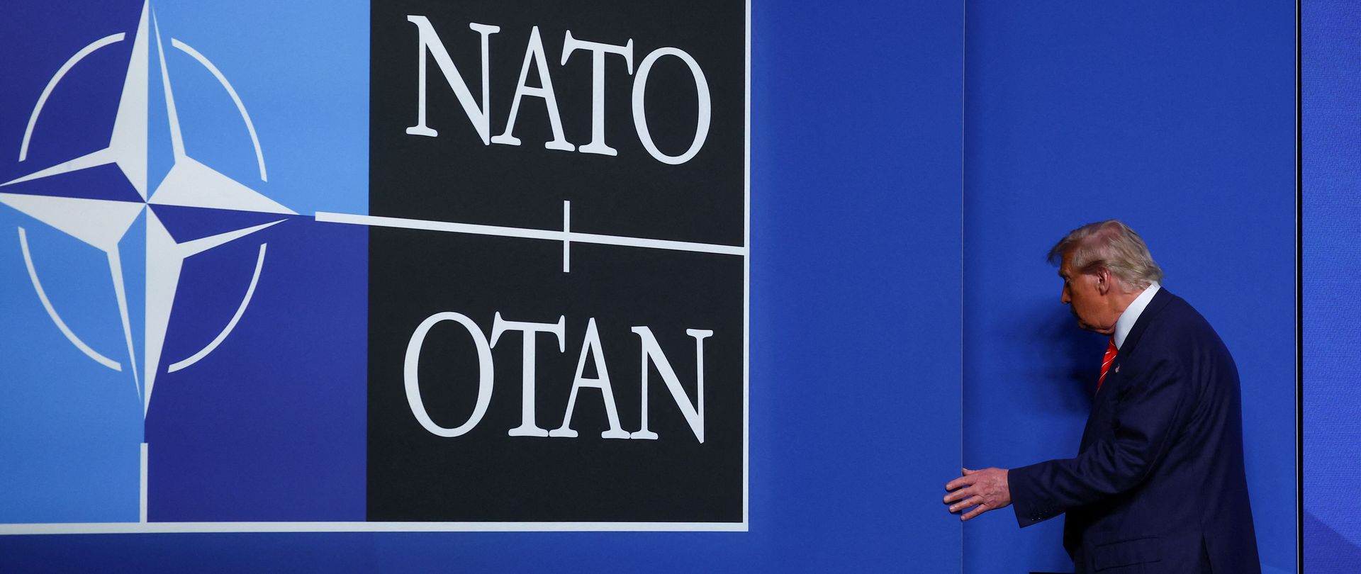 Donald Trump neben einer blauen Wand mit NATO-Logo