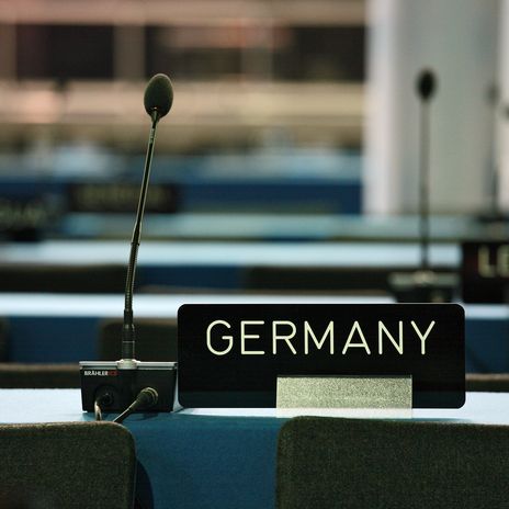 Ein schwarzes Schild mit weißer Aufschrift "Germany" steht in einem leeren Konferenzsaal. Daneben ist ein Mikforon zu sehen.