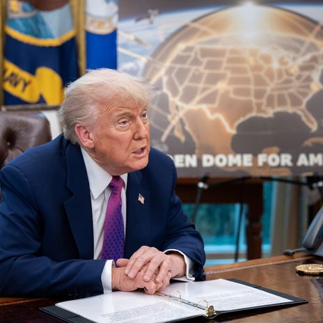 US-Präsident Donald Trump sitzt an seinem Schreibtisch. Im Hintergrund ist ein Poster mit einer goldenen Landkarte der USA zu sehen.
